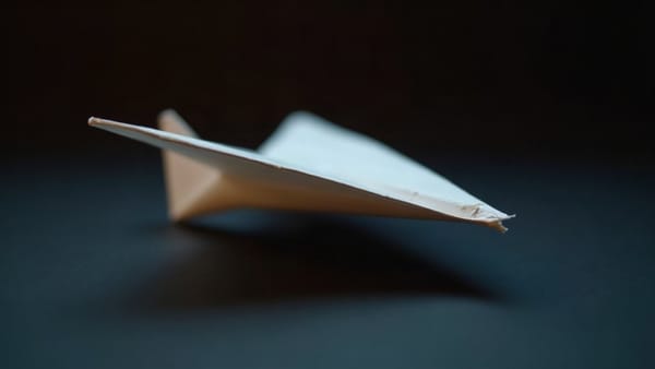 A paper airplane resting on a dark surface with tilt-shift depth of field and warm side-lighting, slightly crumpled at the nose, already in flight and landed.